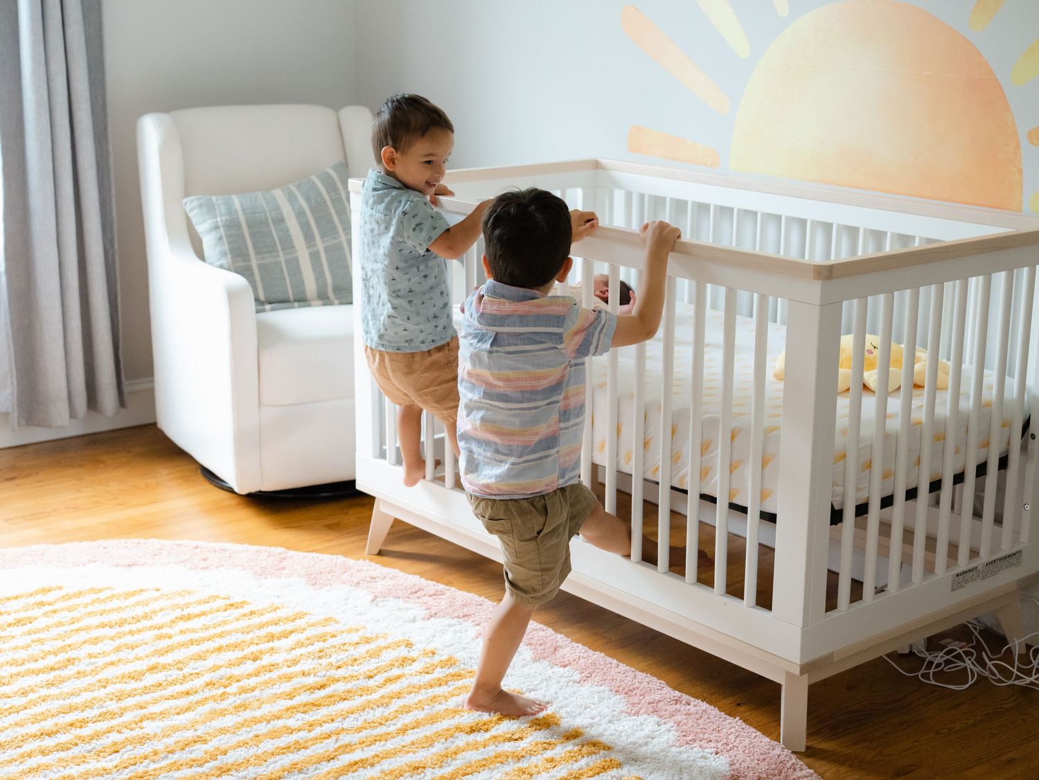 Two brothers jump up on white crib in their sister's nursery with a sun mural on the wall in ocean county nj.