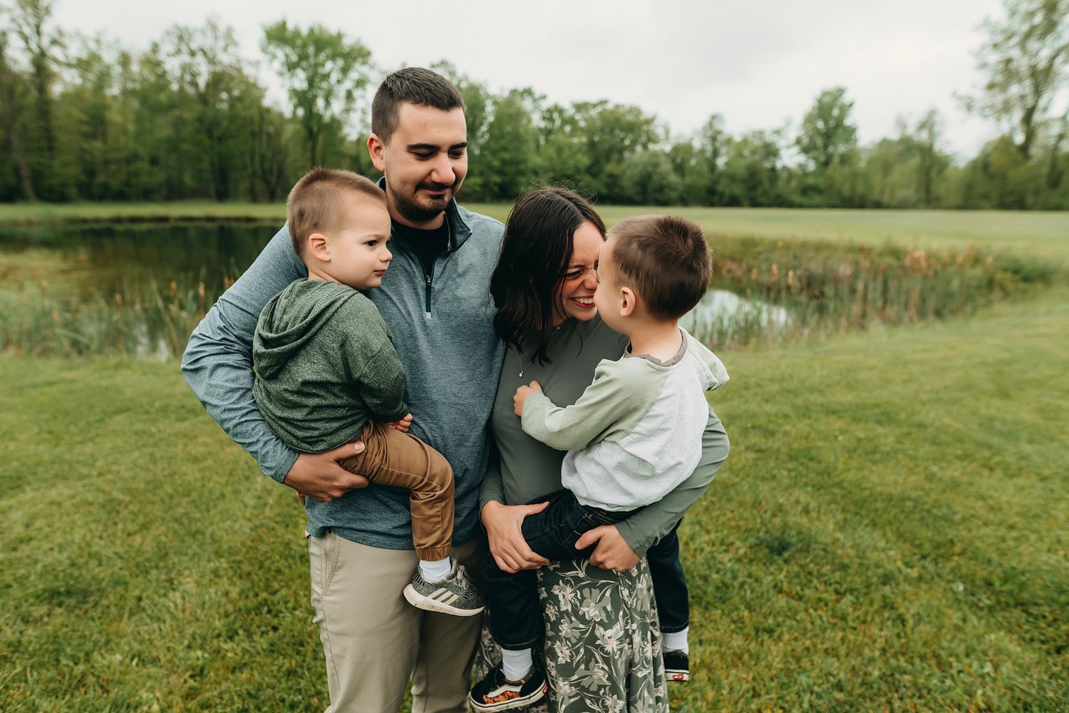 Family of four smiling outdoors near a pond, parents holding young children in a lush green park setting.