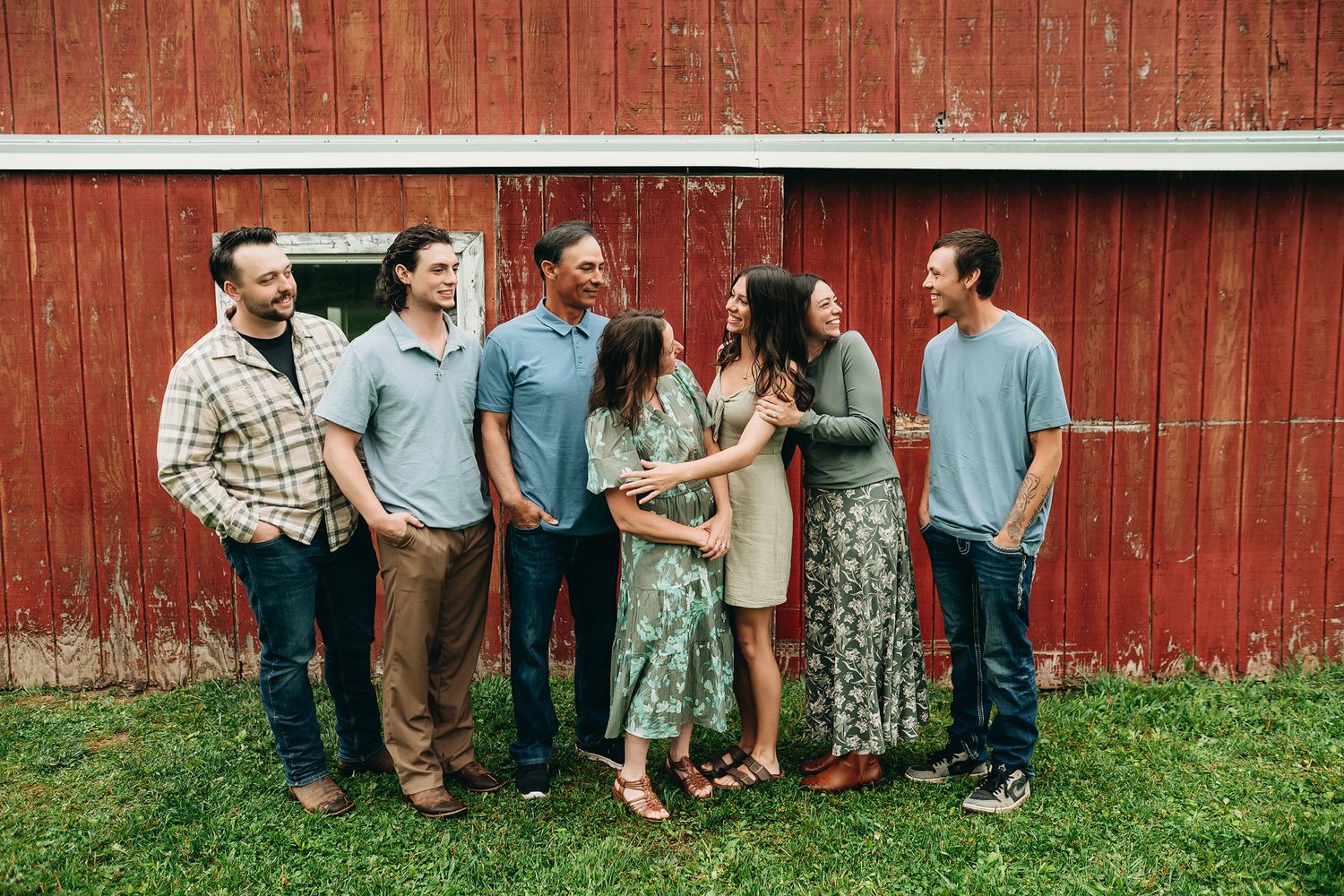 Six adults laughing together in front of a rustic red barn on a green lawn.