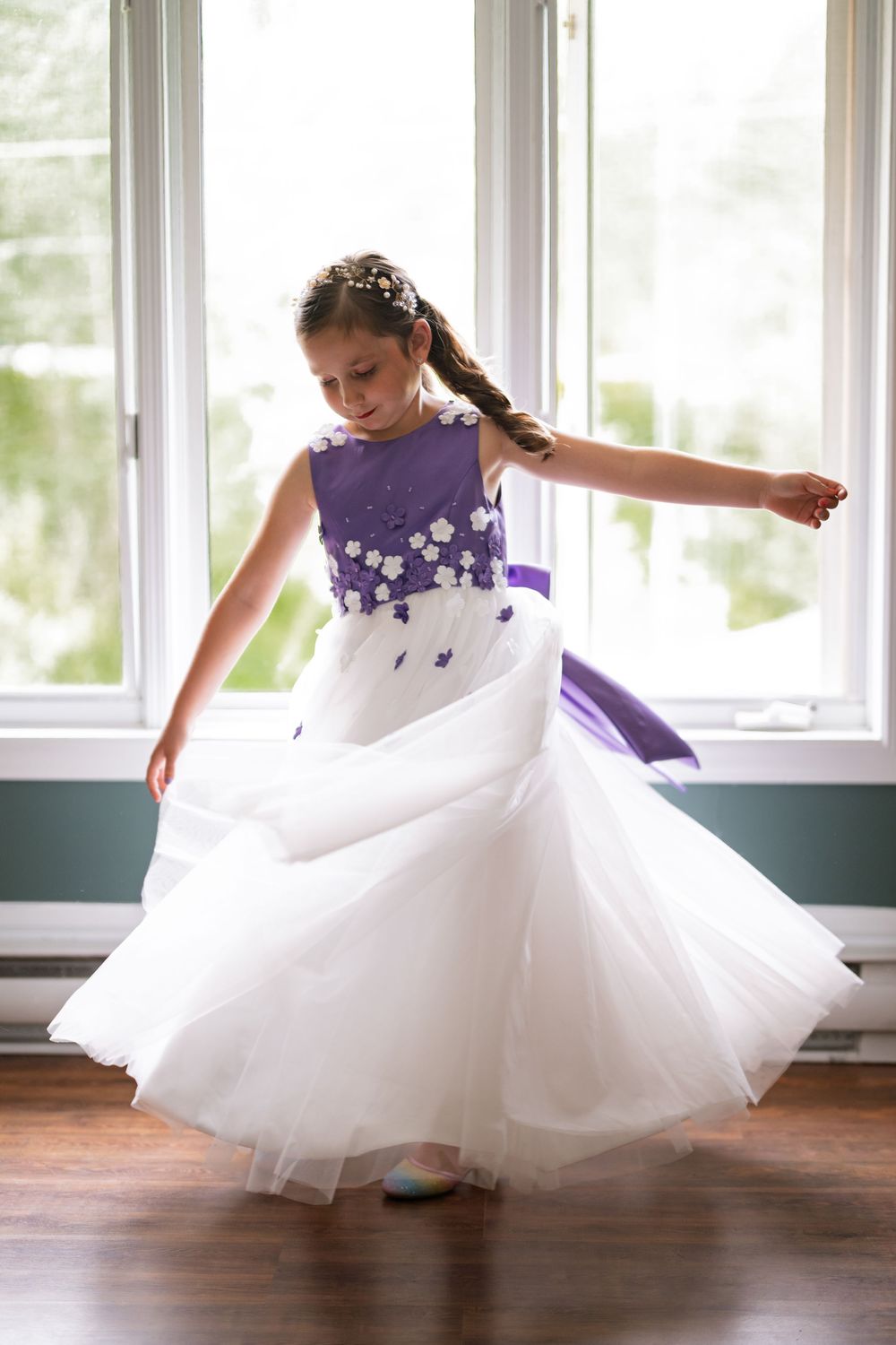 A young flower girl in a white and purple dress twirls gracefully in front of a bright window.