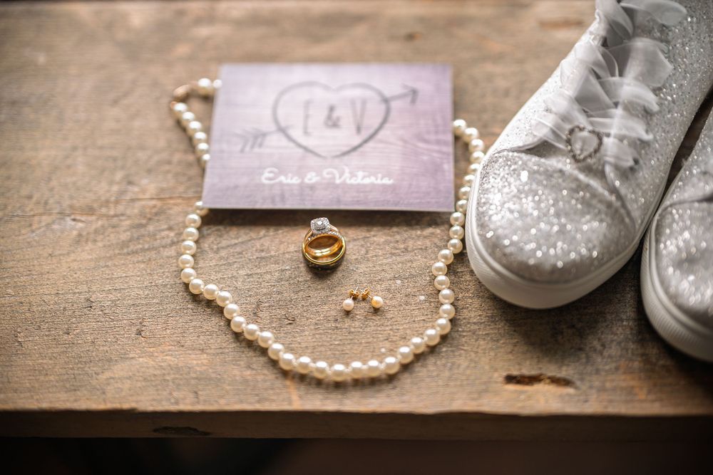 Wedding flat lay with pearl necklace, gold rings, earrings, glitter sneakers, and personalized invitation on rustic wood.
