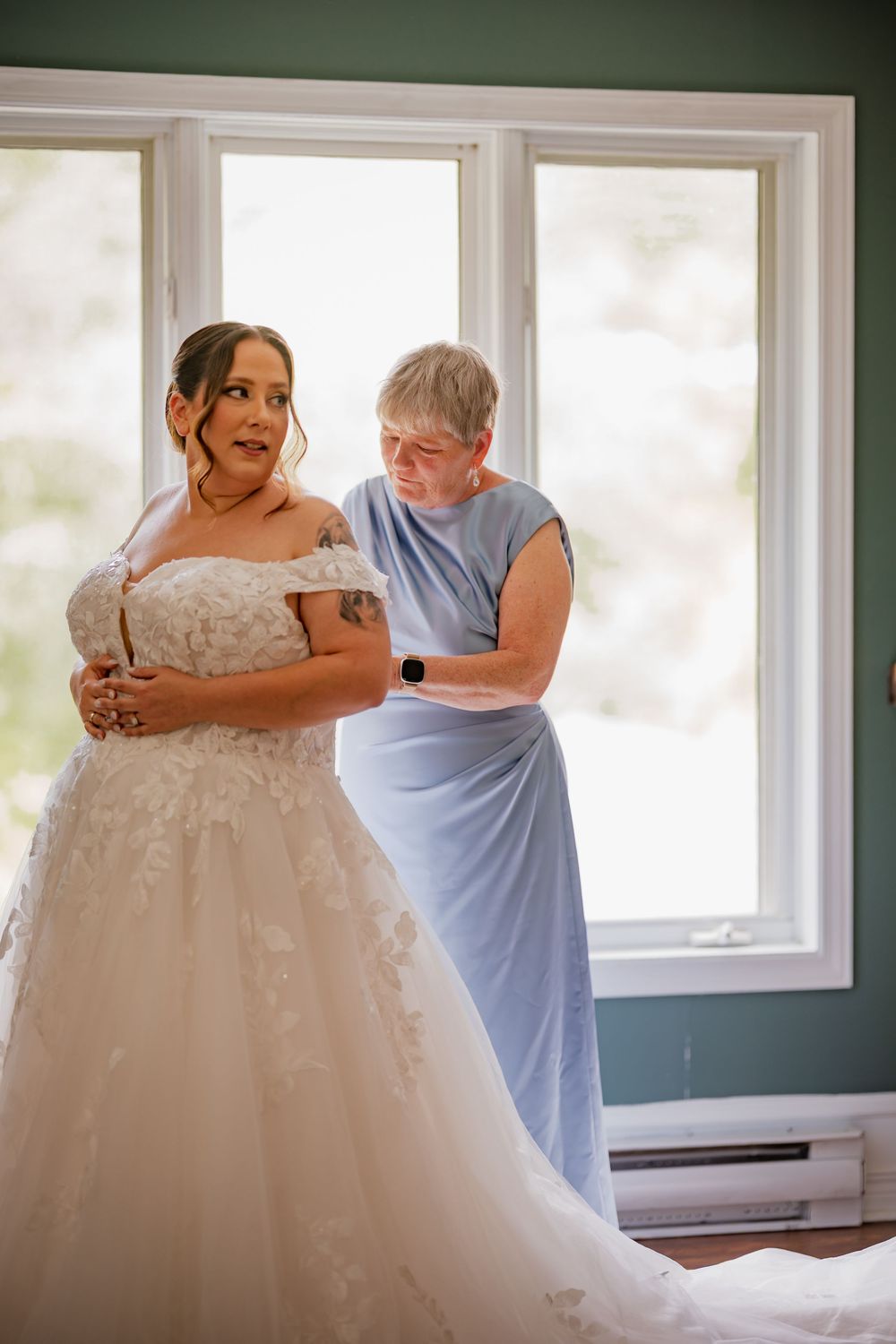 Bride in floral off-shoulder gown being helped by older woman in blue dress near bright window.