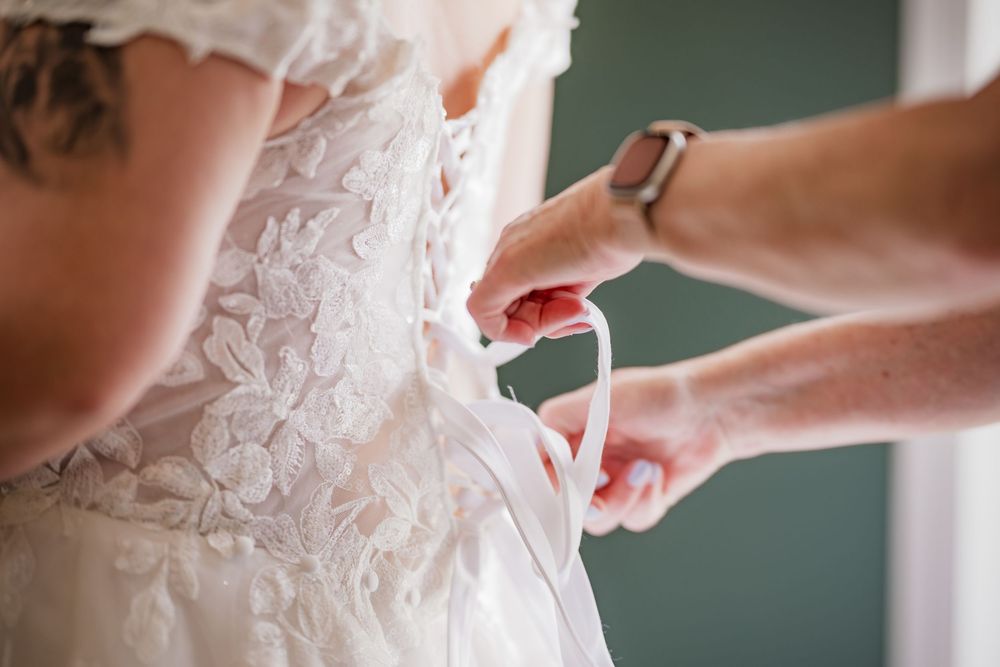 A bride's lace wedding dress being tied at the back by a helper during wedding preparations.