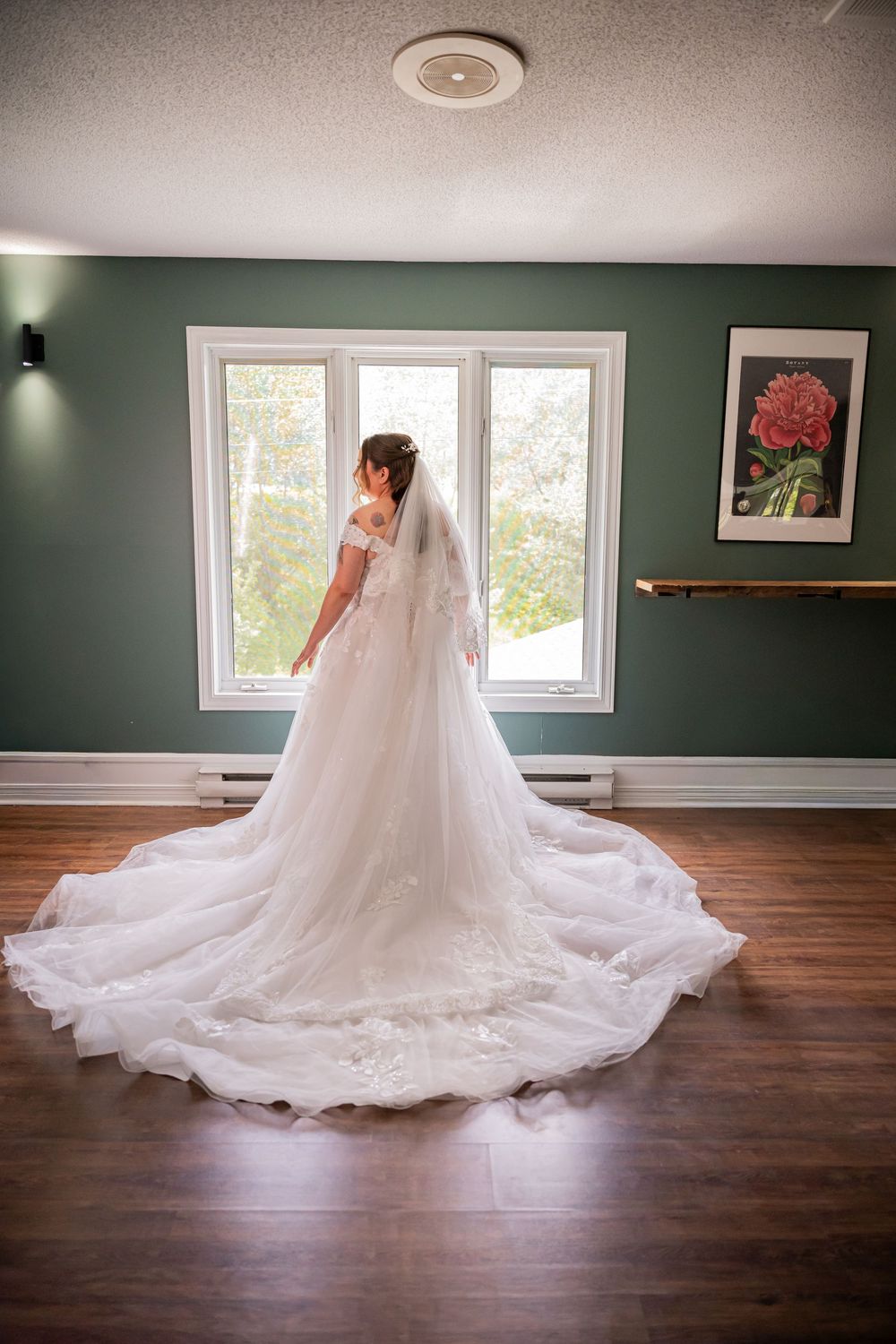 Bride in elegant white lace wedding gown with long train and veil stands by window in green room.