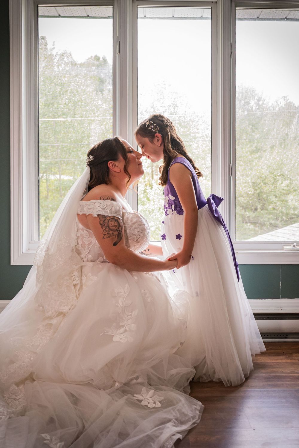 Bride in white lace gown shares tender moment with flower girl in purple dress by sunlit window.
