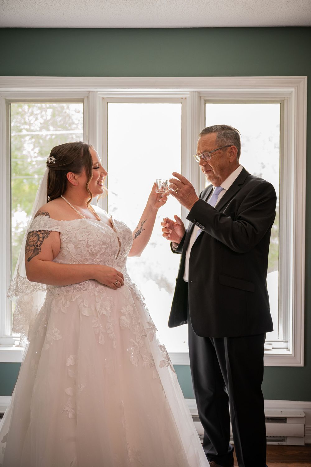 Bride in lace gown toasts champagne with older man in tuxedo by bright window.