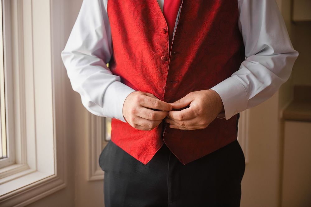 Man in white dress shirt buttoning a red satin vest, preparing for a formal event near a window.