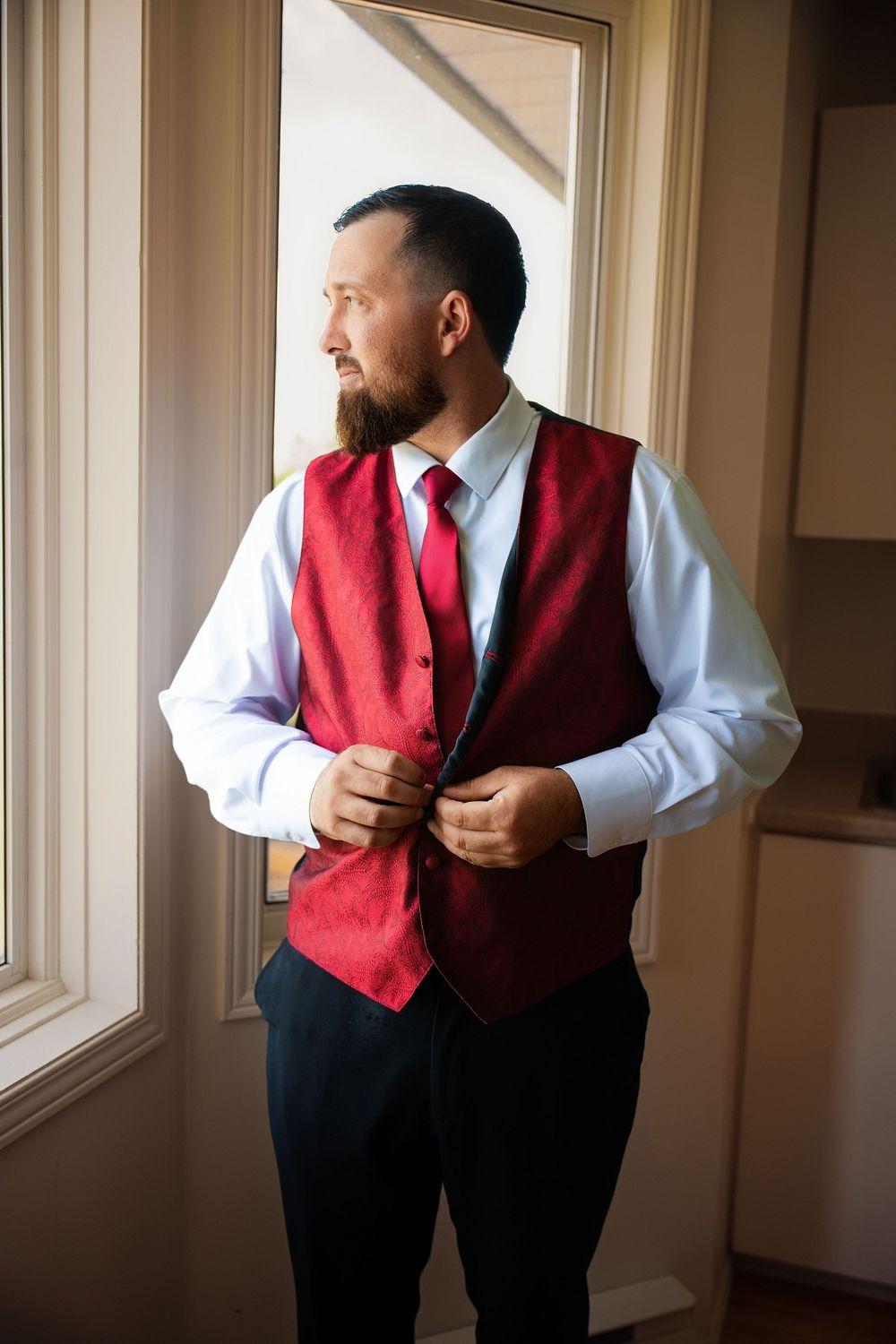 Bearded groom buttoning red velvet vest with matching tie while standing near window.