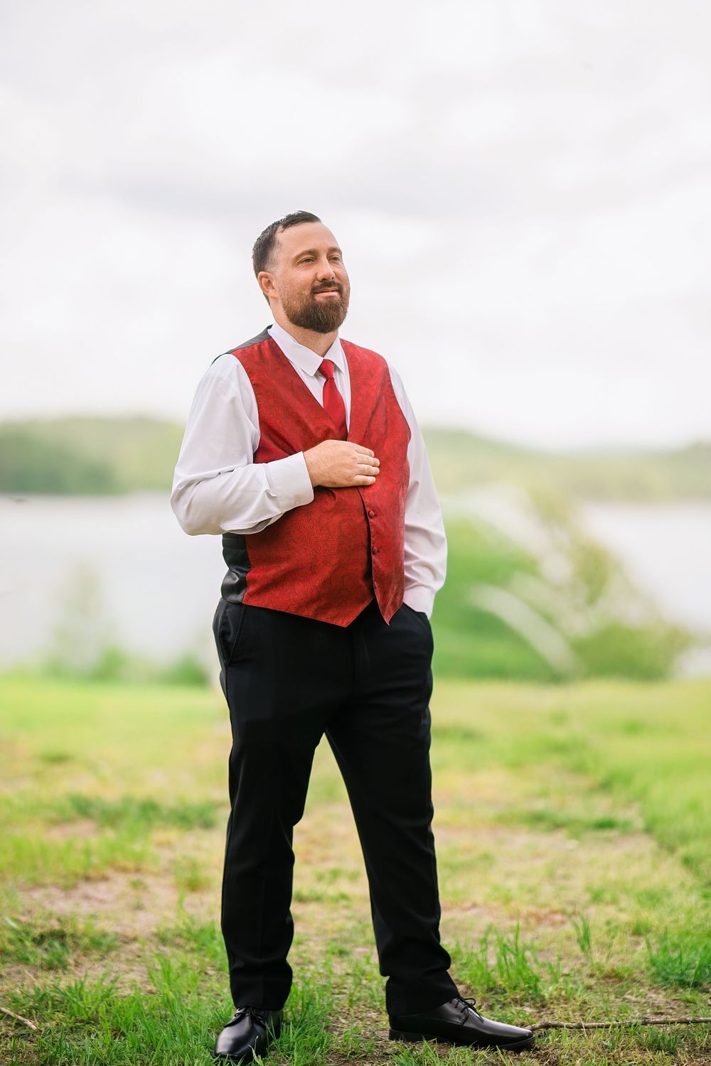 Bearded groom in red vest and tie poses outdoors near a lake on his wedding day.