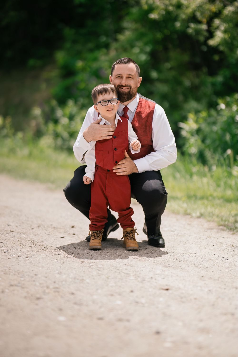 Smiling bearded father crouching behind young son in red suit on a sunny outdoor path.