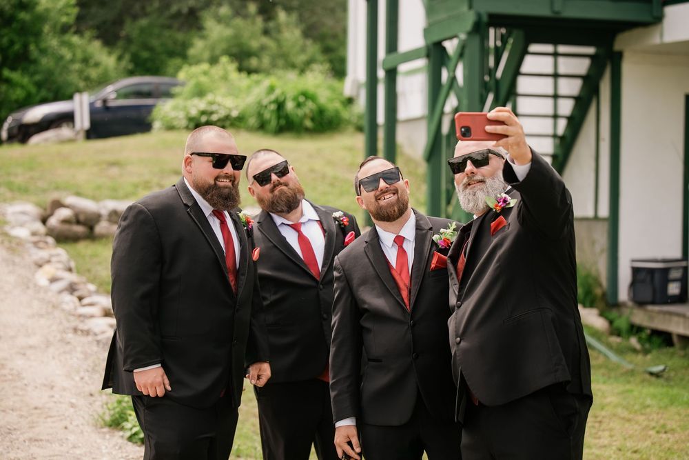 Four groomsmen in black suits and red ties take a selfie outdoors at a wedding venue.