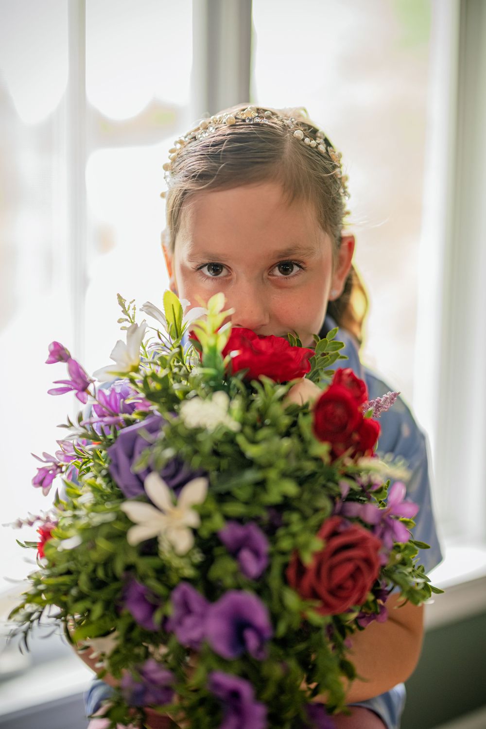 Young girl smelling a colorful bouquet of red, purple, and white flowers near a bright window.
