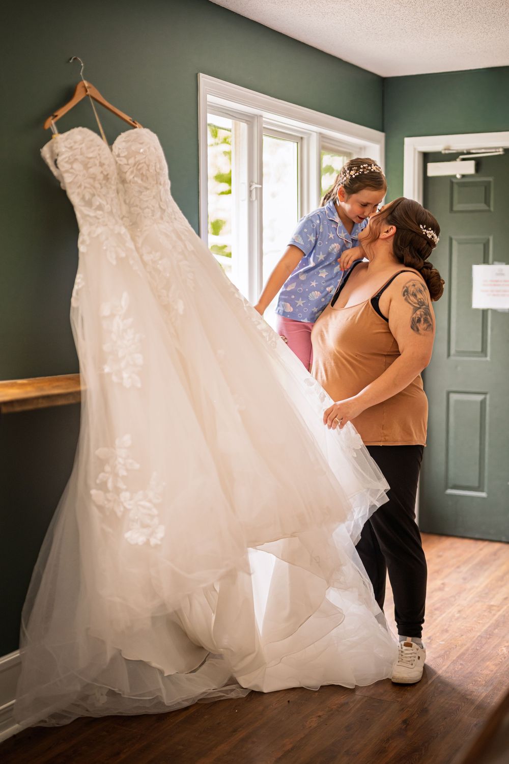 A bride-to-be shares a tender moment with a young girl beside a hanging lace wedding gown in a green-walled room.