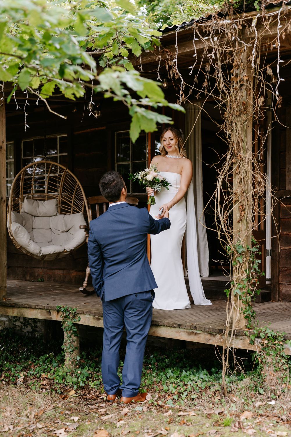 Groom in navy suit presents bouquet to smiling bride in white gown on rustic wooden porch with hanging chair.