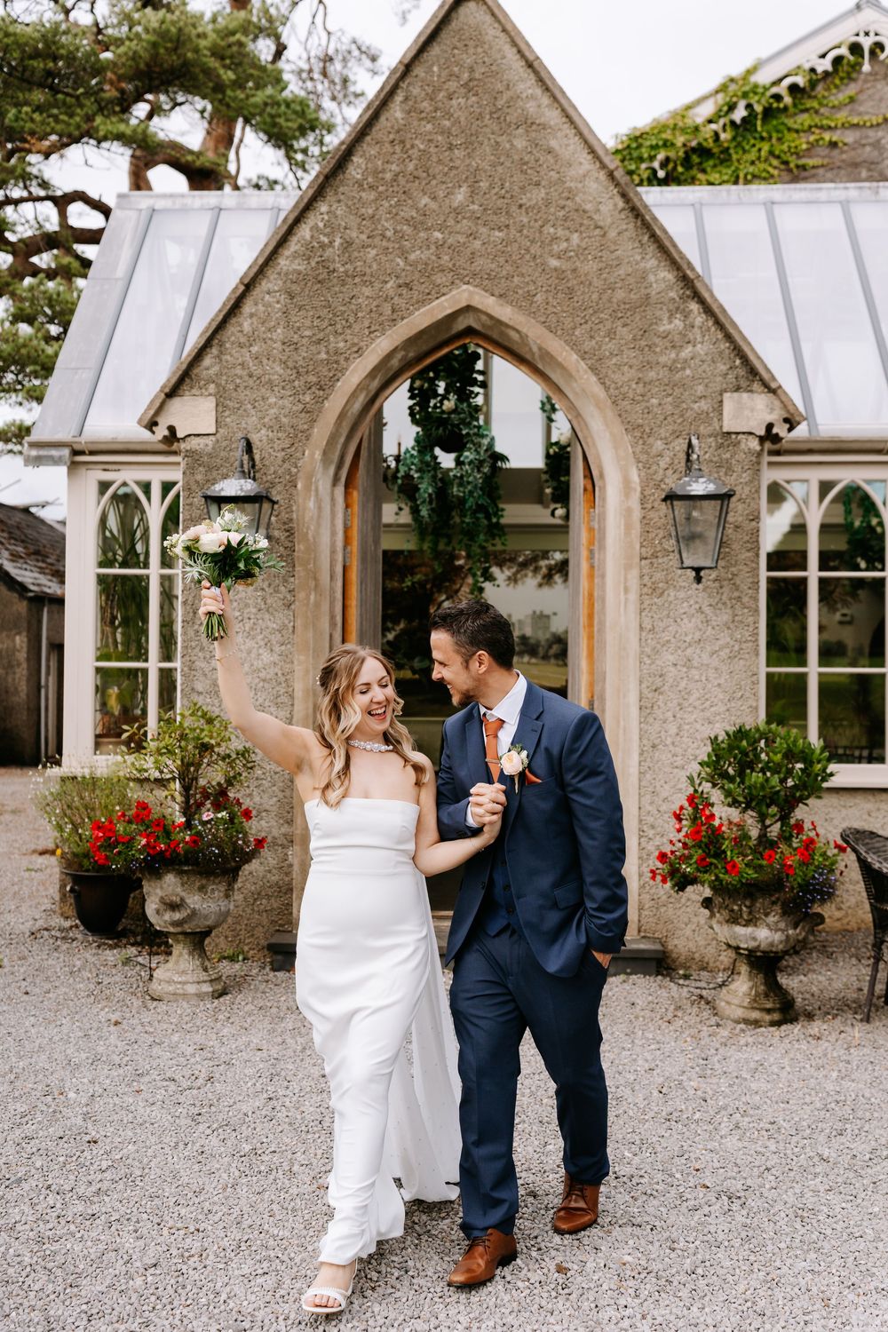 Joyful newlyweds celebrate outside a stone chapel, bride raising bouquet in white gown, groom in navy suit with coral tie.