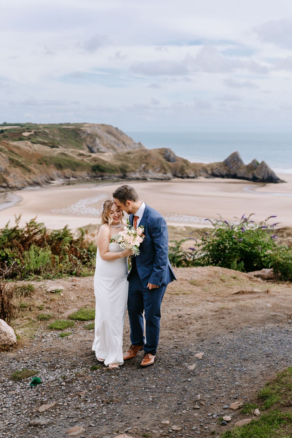 A bride and groom share a tender moment on a coastal clifftop path, with a stunning sandy bay and dramatic cliffs behind them.