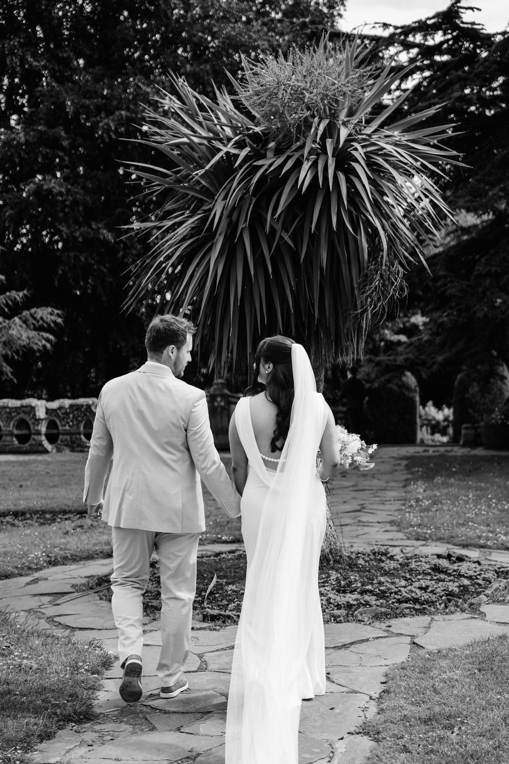 Black and white photo of a bride and groom holding hands walking through a lush garden on their wedding day.