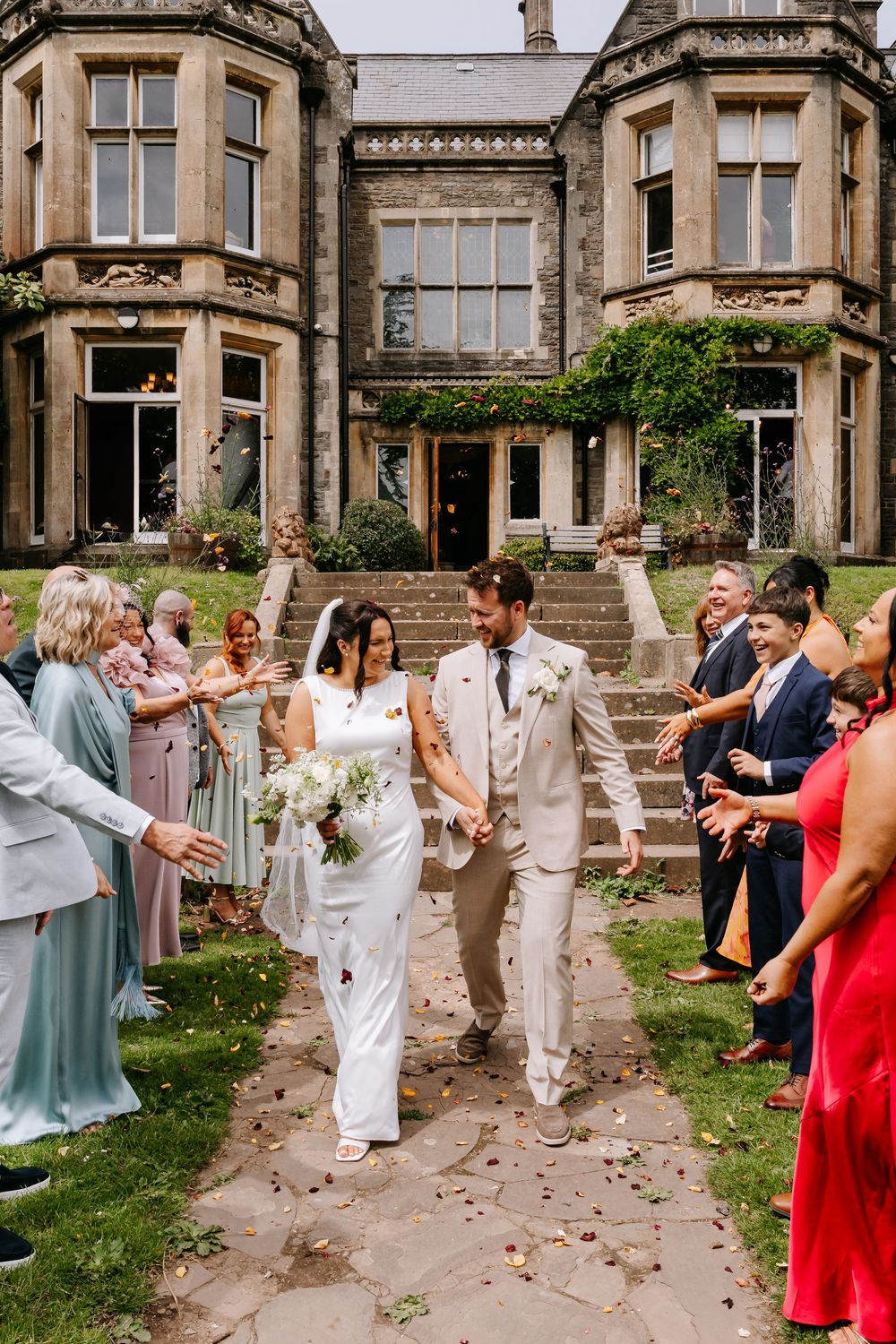 Bride and groom walk through confetti outside a grand stone manor house, surrounded by smiling wedding guests.