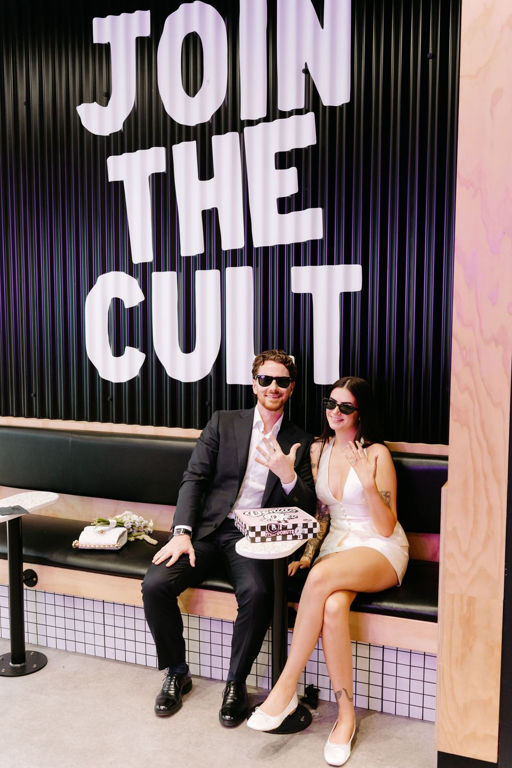 Couple in formal wear sitting on bench under Join The Cult sign, holding checkered cake, wearing sunglasses.