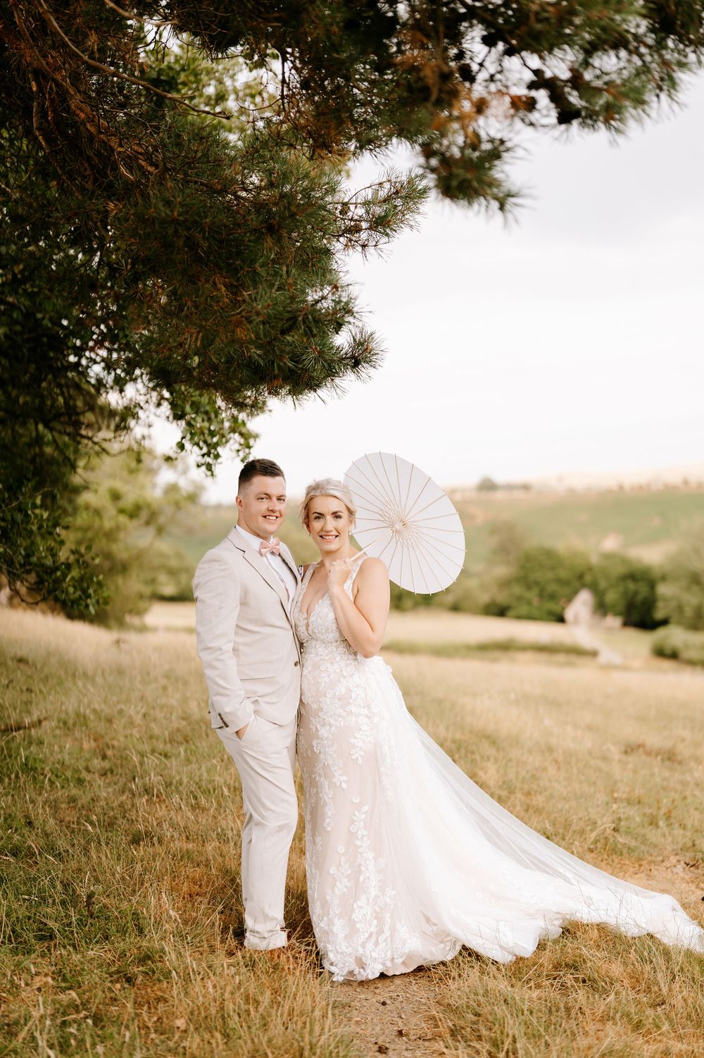 Bride and groom pose under pine tree in lush field, bride holds white parasol wearing lace gown, groom in cream suit.