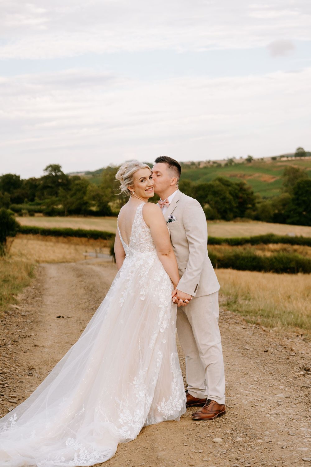 Bride and groom holding hands on a rural dirt path, bride in lace gown, groom in tan suit, rolling green fields behind.