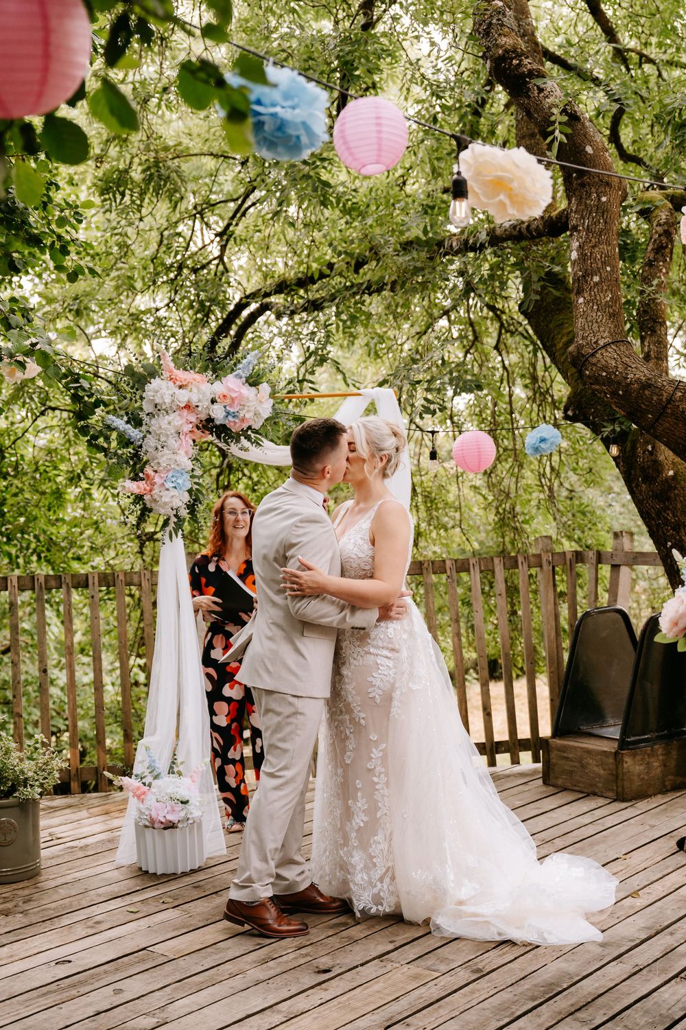 Bride and groom share first kiss on outdoor deck wedding ceremony with floral arch and colorful paper lanterns.