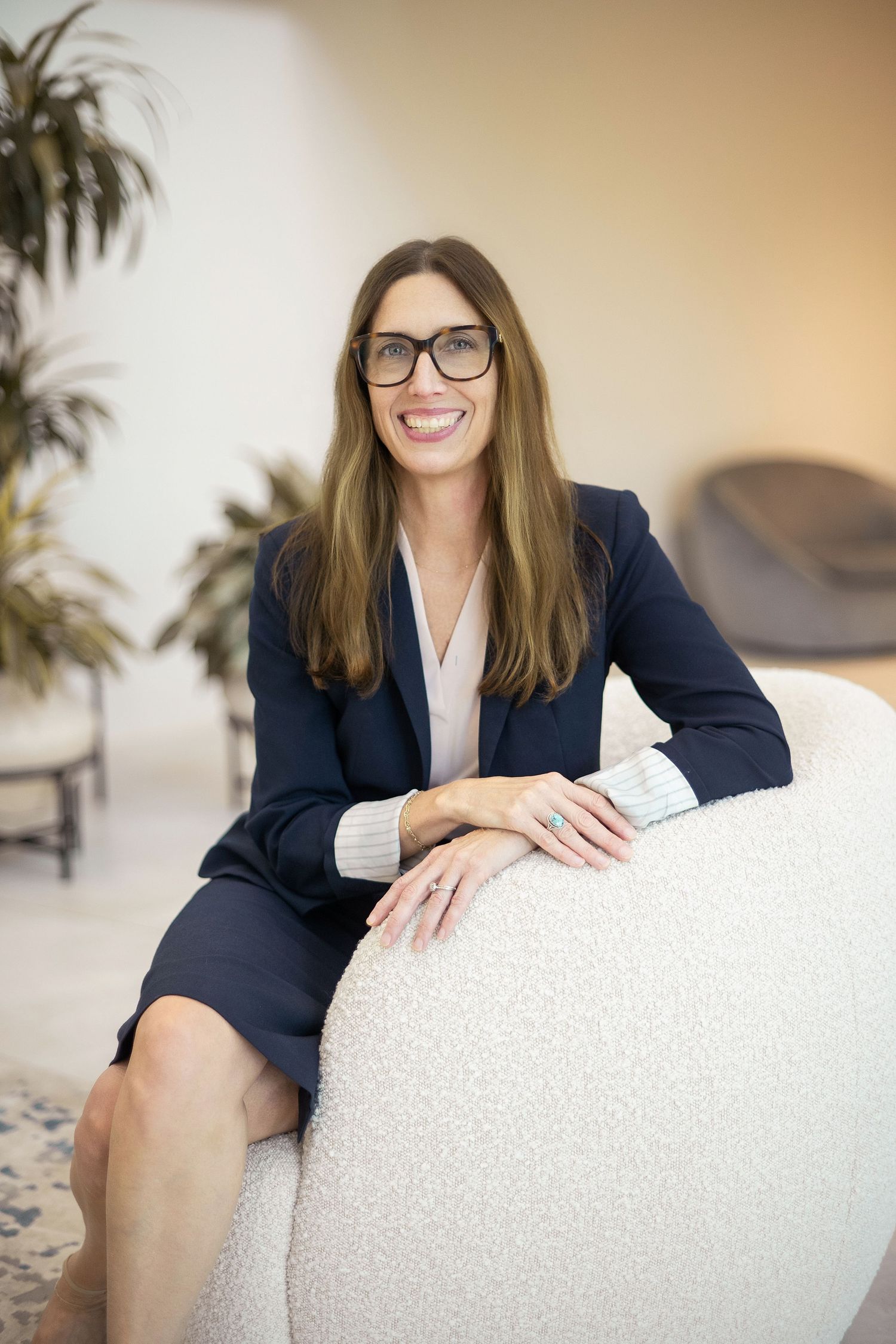 Smiling businesswoman in navy blazer and glasses sitting relaxed on white chair in modern office having a headshot taken.