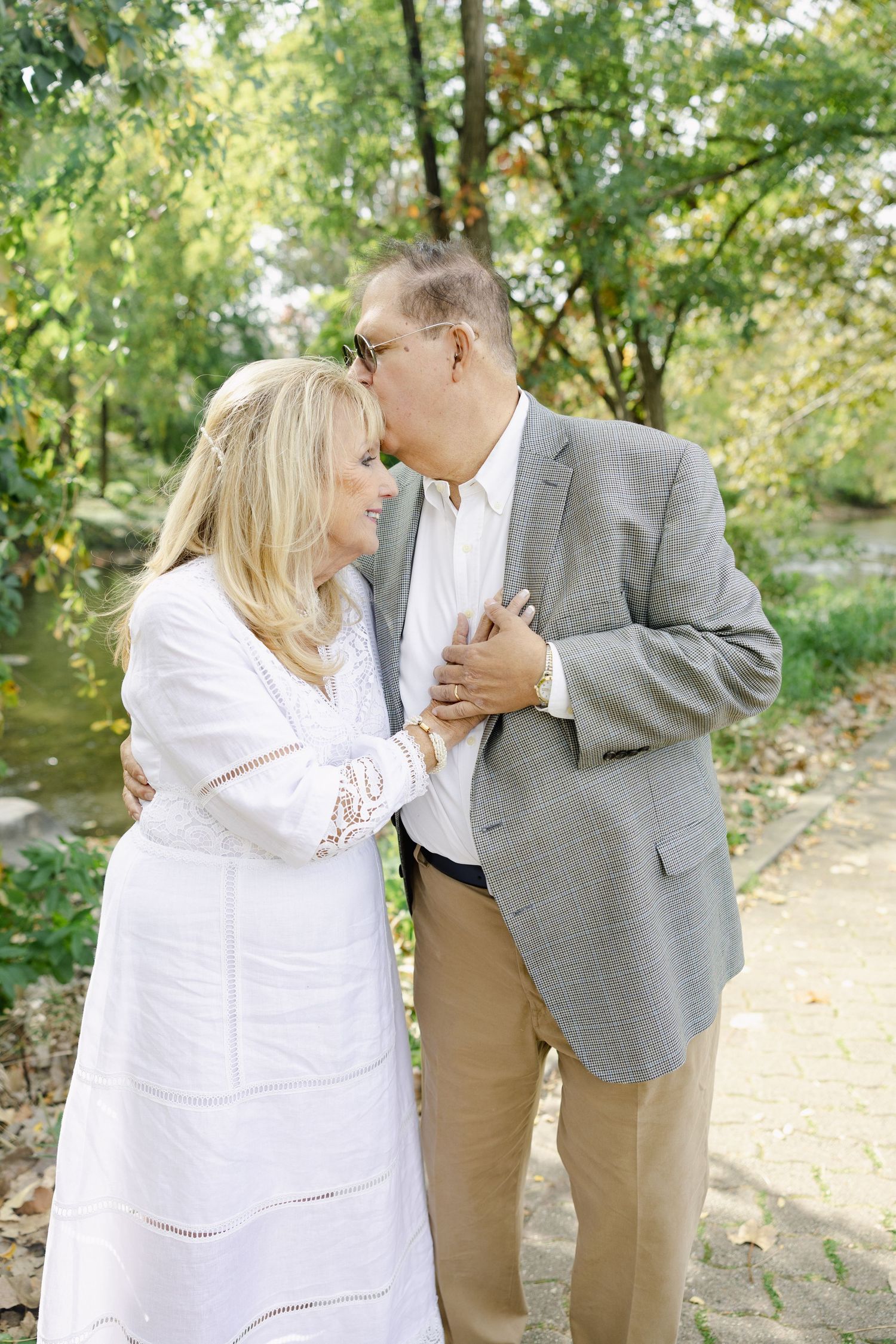 Older couple sharing a tender kiss outdoors on a scenic tree-lined path near a stream.