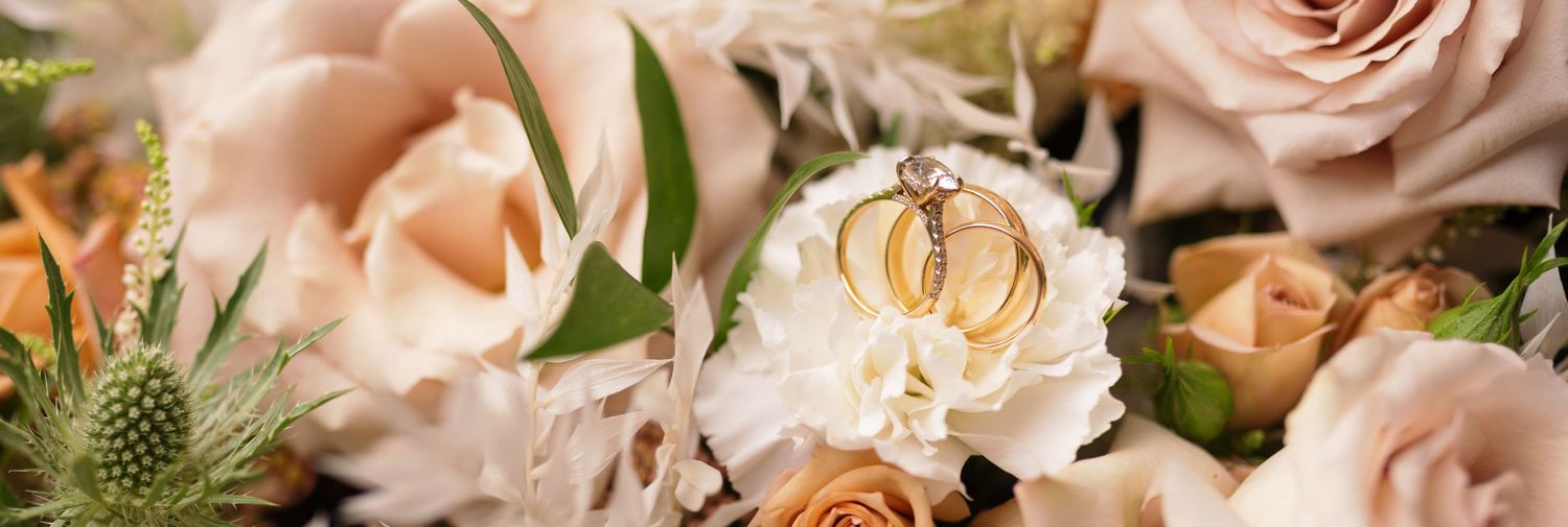 Close-up of two gold wedding rings resting on white carnation blooms surrounded by blush pink roses and greenery.