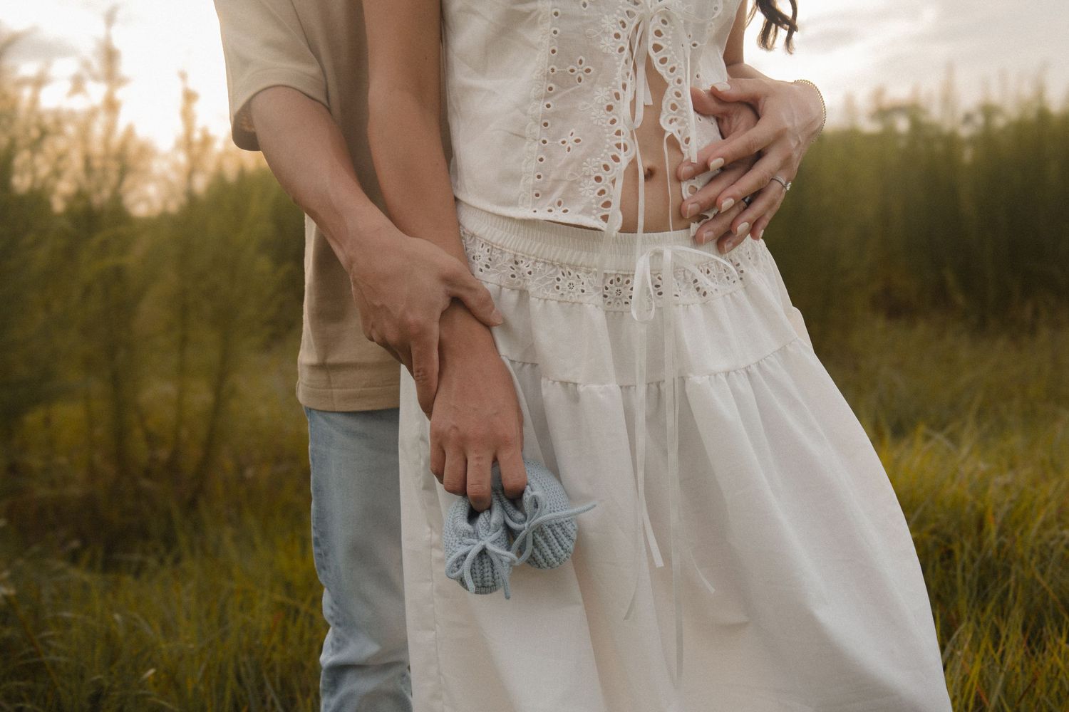 Couple holding tiny baby booties outdoors in a field, announcing their pregnancy at sunset.