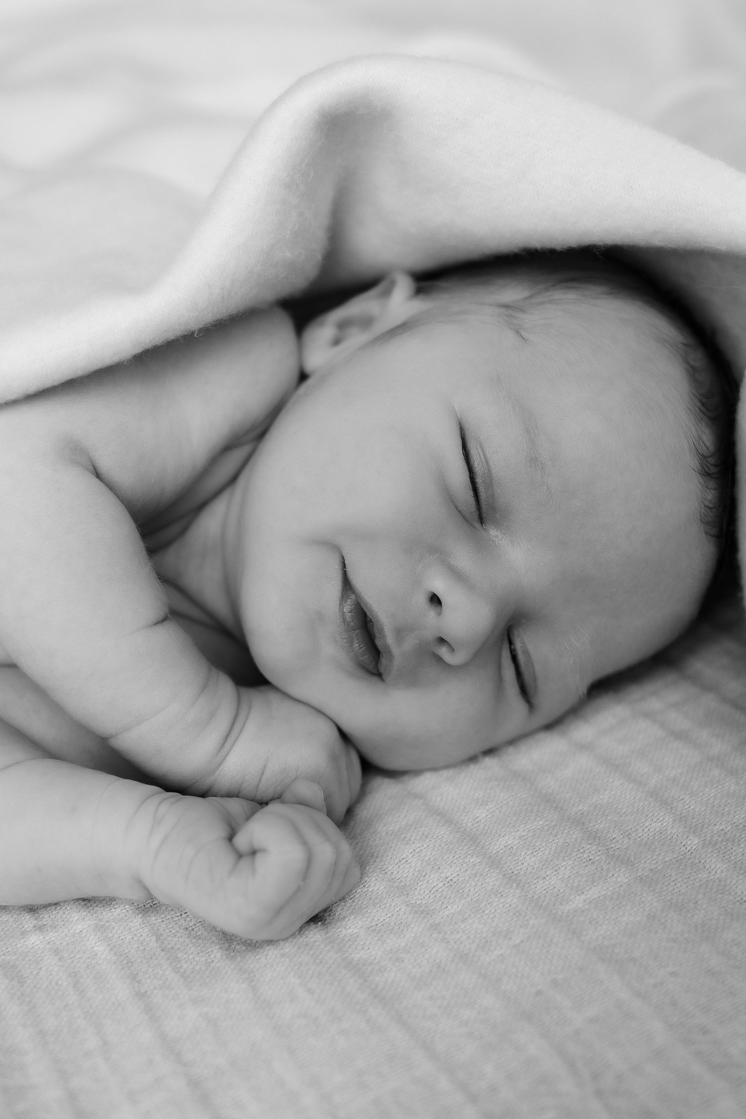 Black and white photo of a peaceful sleeping newborn baby with tiny fists, resting under a soft white blanket.