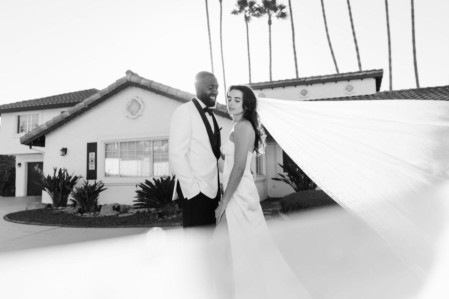 Black and white wedding photo of bride and groom embracing outdoors near a building with palm trees.