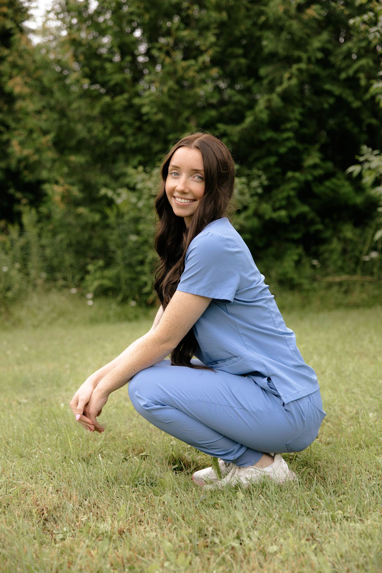Young woman in blue scrubs crouching on grass, smiling outdoors with green trees in background.