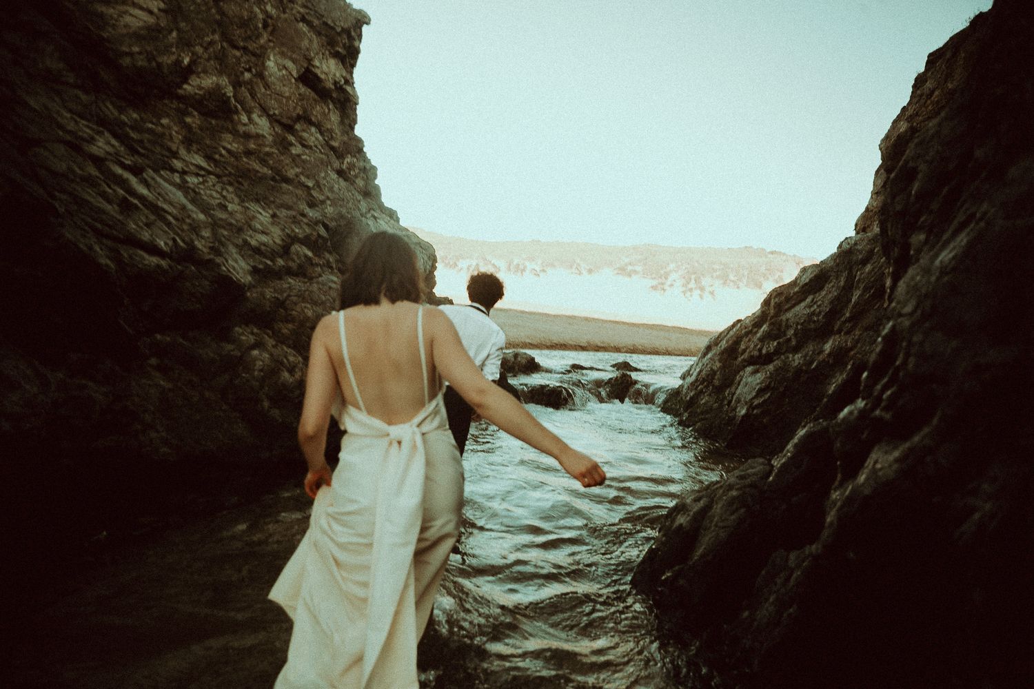 Couple in white dress walking between rocky coastal cliffs toward the ocean waves.