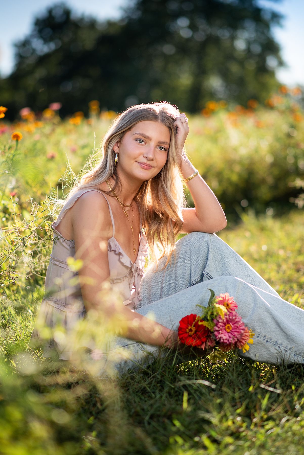 Young blonde woman sitting in a sunny flower field wearing gray jeans, holding colorful zinnia blooms.
