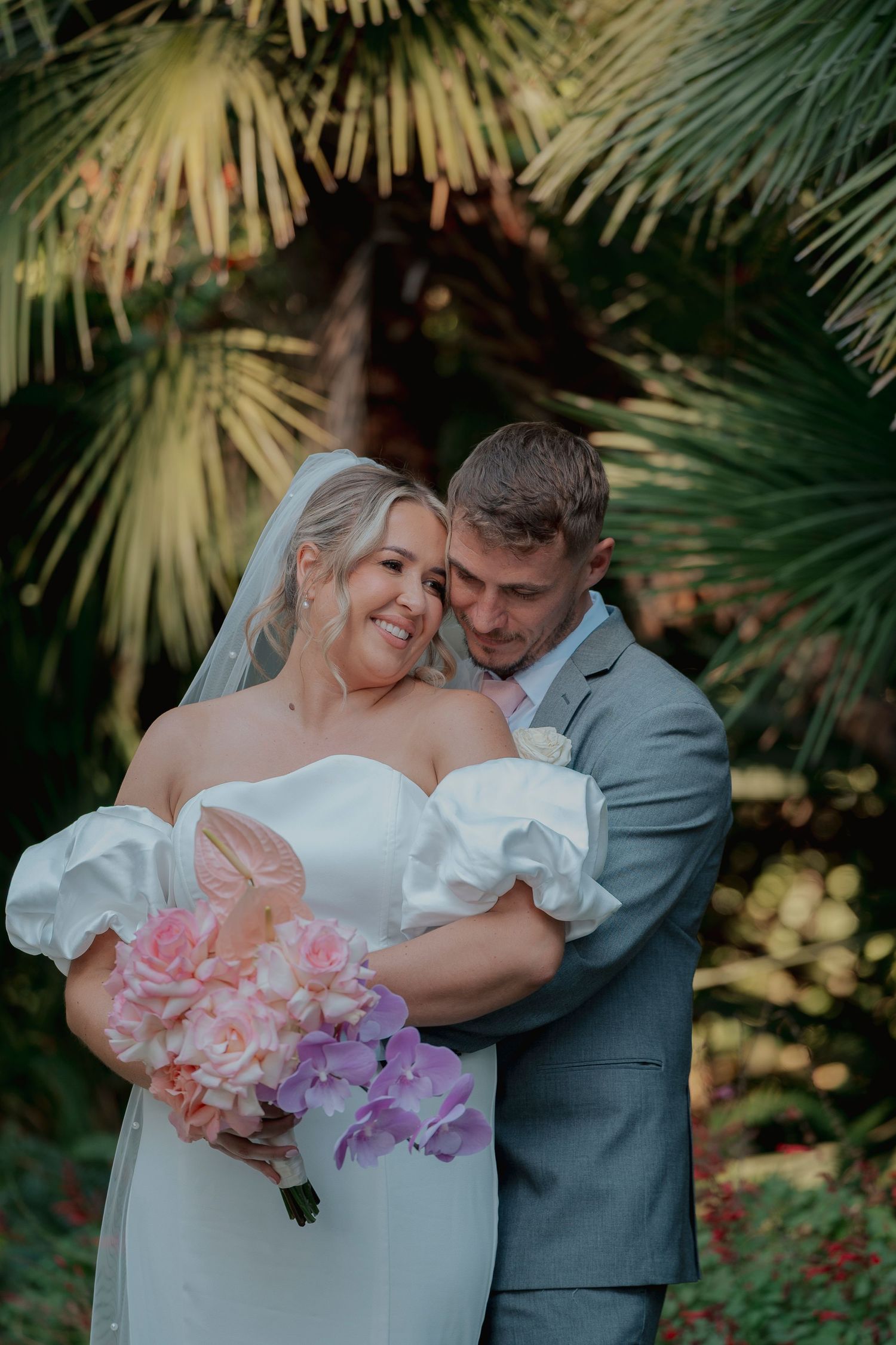 A bride and groom share a tender moment, holding hands near a decorative window, groom in dark suit with boutonniere.