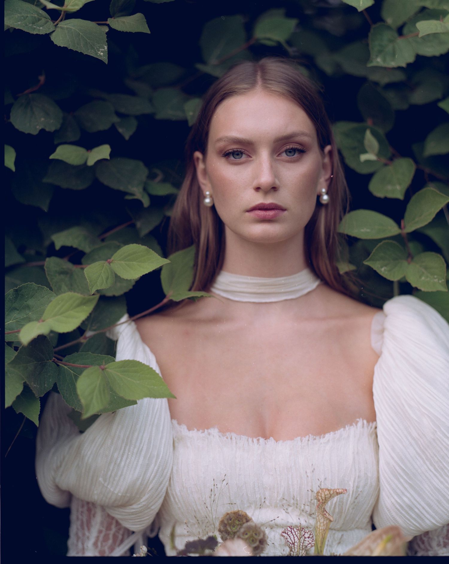 A young woman in a white ruffled dress with pearl earrings and choker poses among lush green leaves.