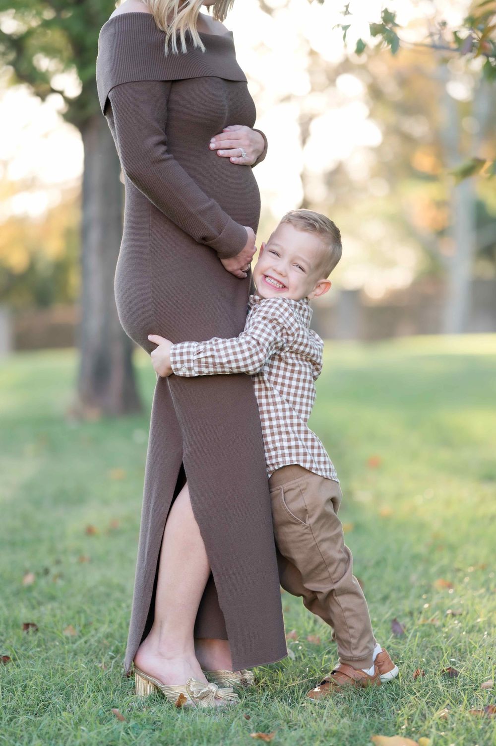 A young boy hugs his pregnant mother's belly outdoors during a maternity photo session in a park.