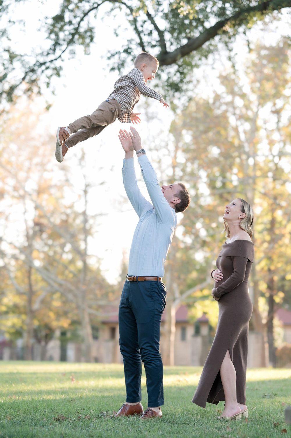 A father joyfully tosses his toddler in the air while his pregnant wife laughs beside him in a sunny park.