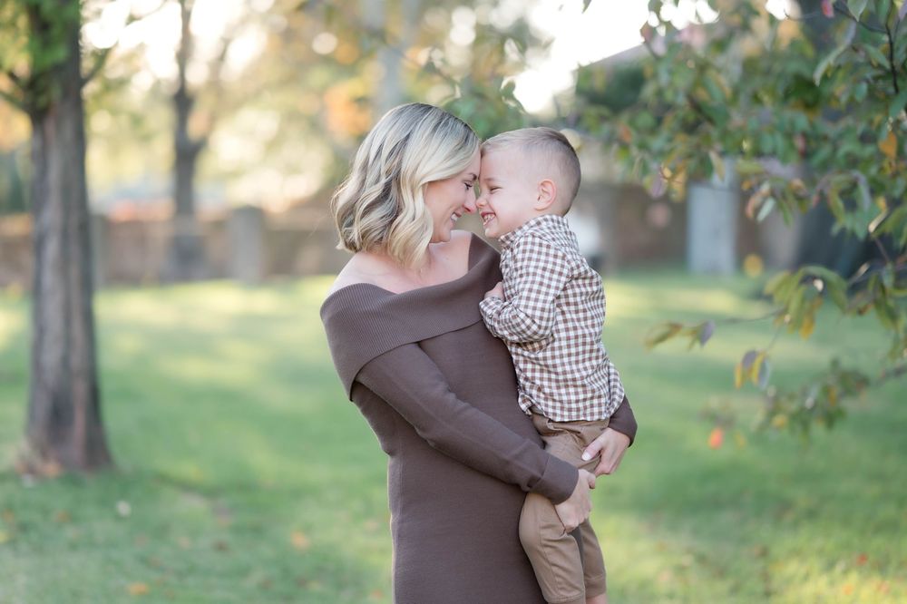 A blonde woman in a brown off-shoulder dress holds and nuzzles a smiling toddler boy outdoors in a sunny green park.