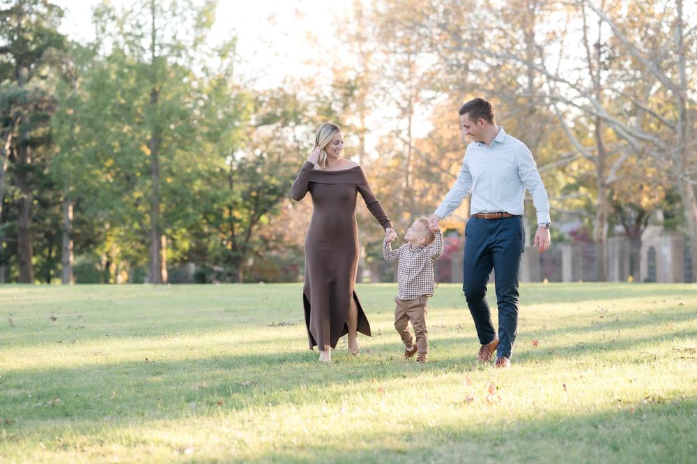A happy family of three walks hand-in-hand through a sunny green park, parents swinging their toddler between them.