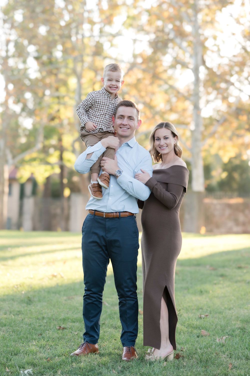 A smiling family of three poses outdoors in fall, dad holding toddler son, mom in brown maternity dress.