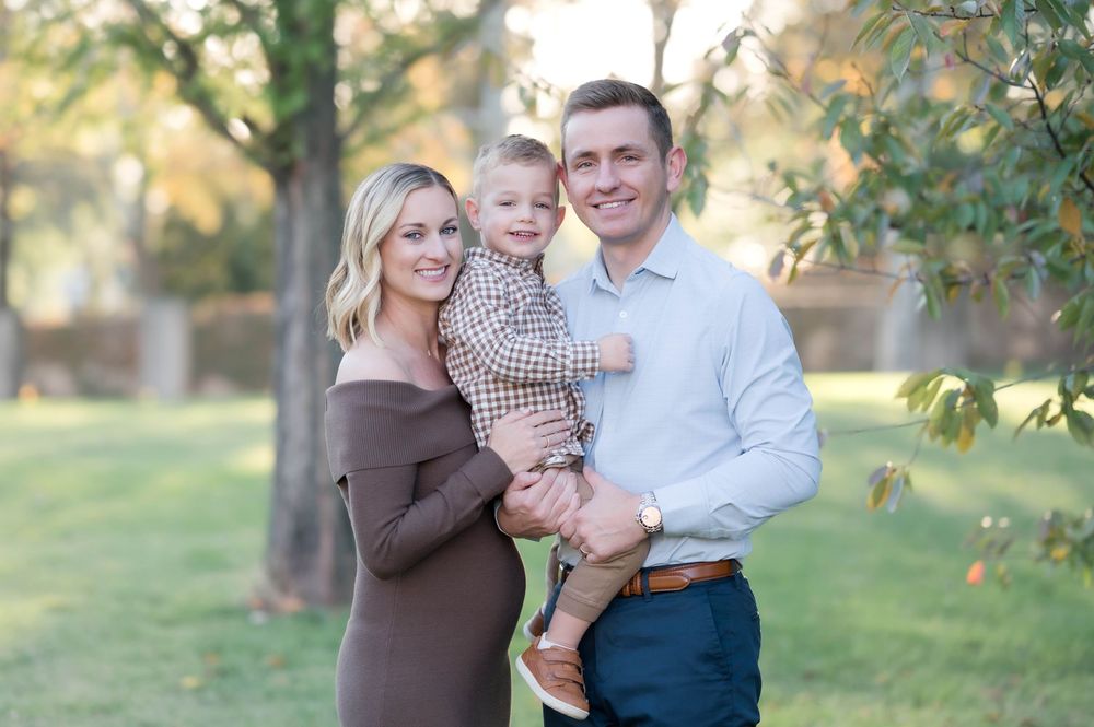 A smiling family of three poses outdoors in a park, with a woman in brown, a man in white, holding a toddler.
