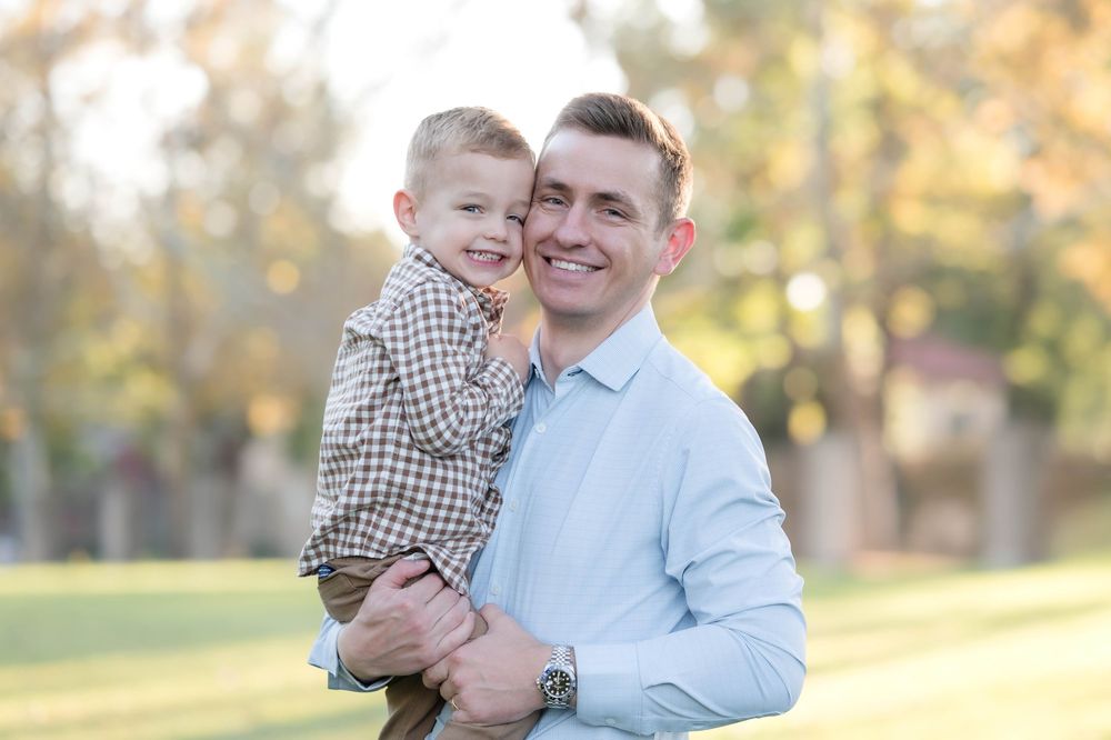 A smiling man holds a young boy in a plaid shirt outdoors in a sunny park with green trees in the background.
