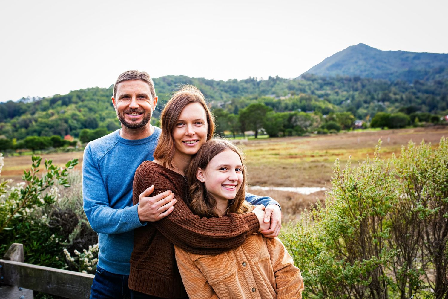 A happy family of three poses outdoors with lush green hills and a scenic valley in the background.