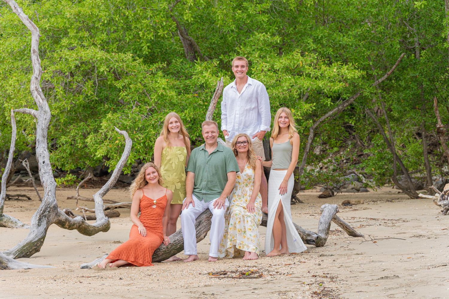 Family of six posing on a sandy beach surrounded by driftwood and lush green tropical trees.