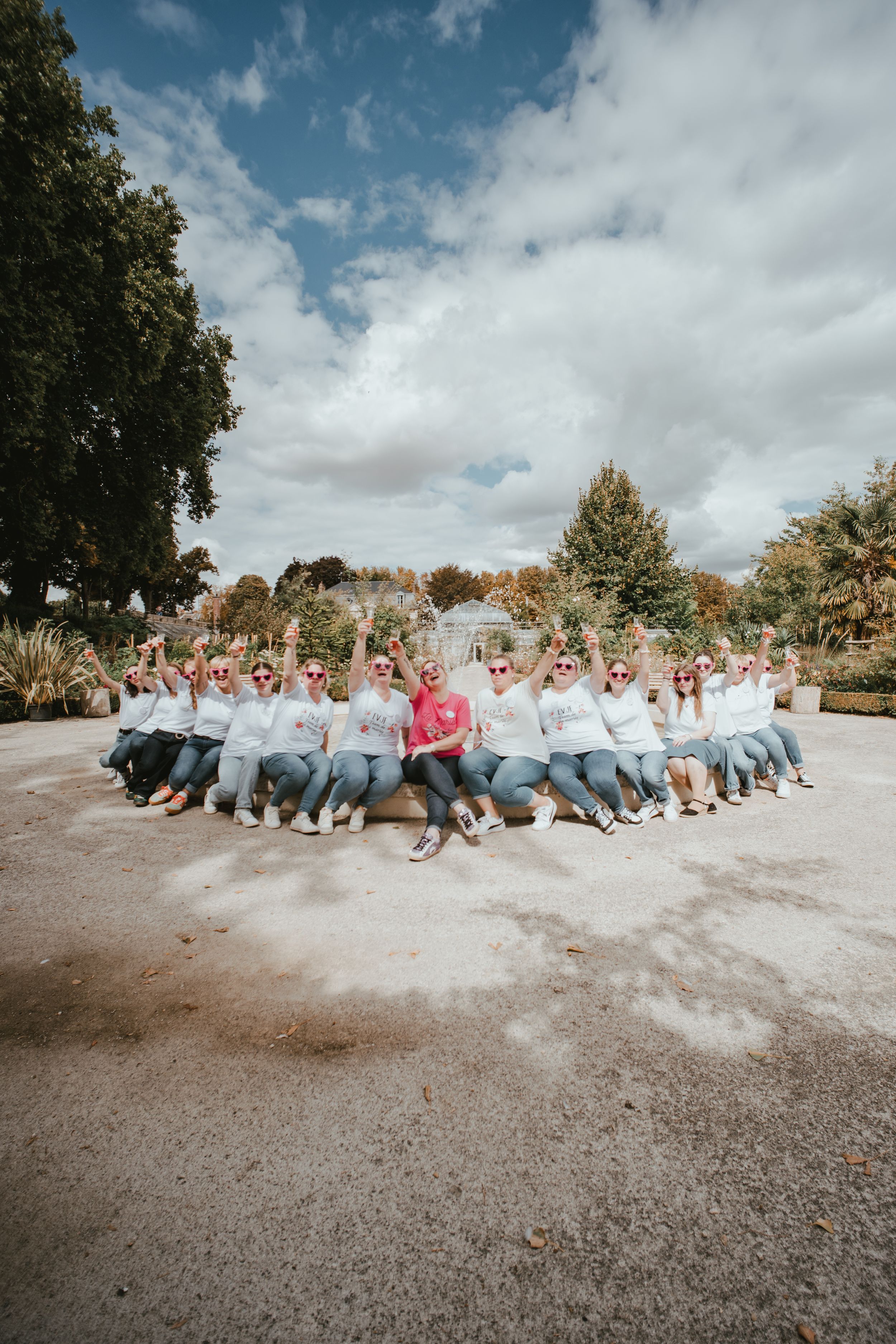 Large group of women in white hoodies sitting together outdoors, raising hands joyfully in a scenic park setting.