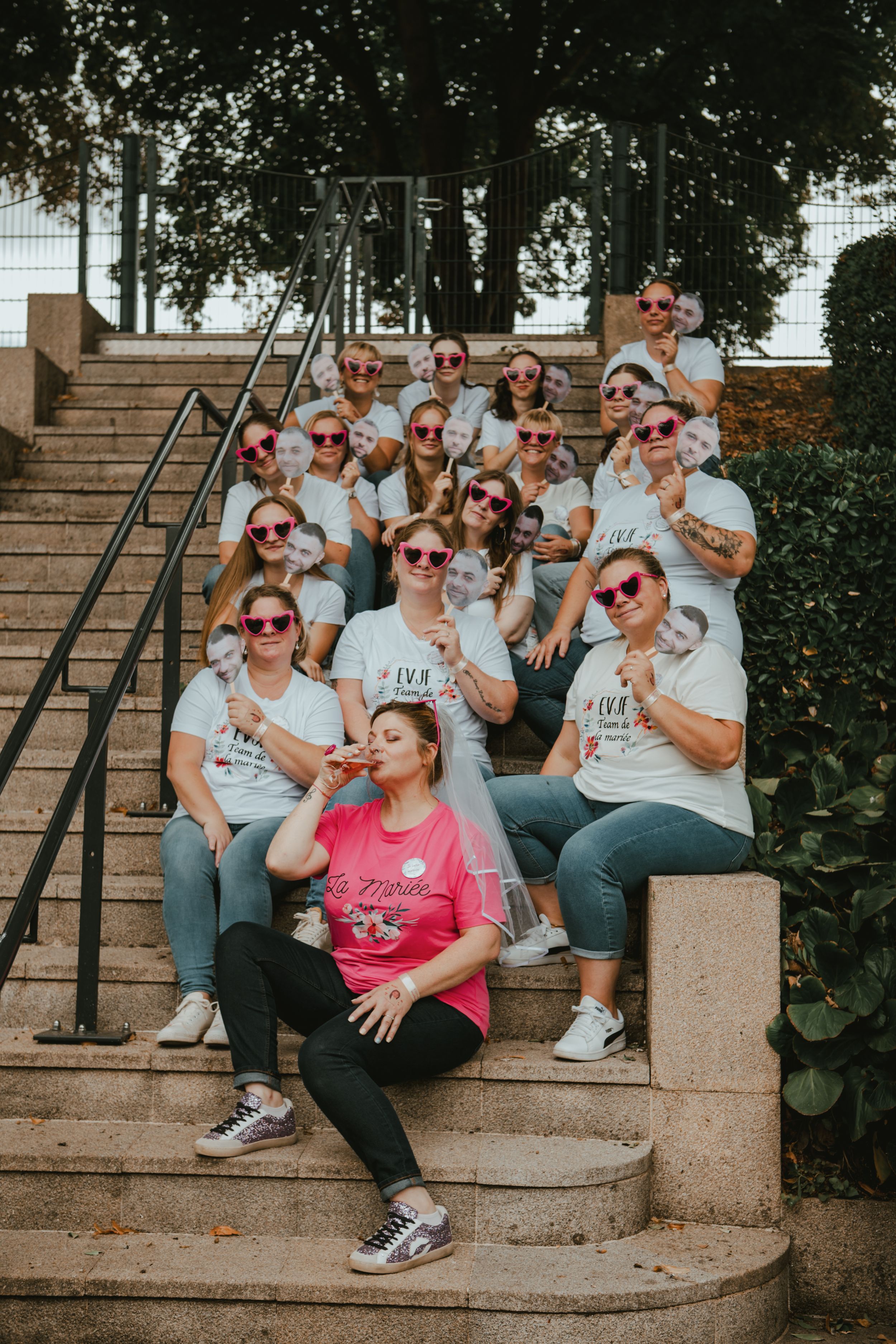 Large bachelorette party group on outdoor steps, wearing matching white shirts and sunglasses, bride in pink holding champagne.
