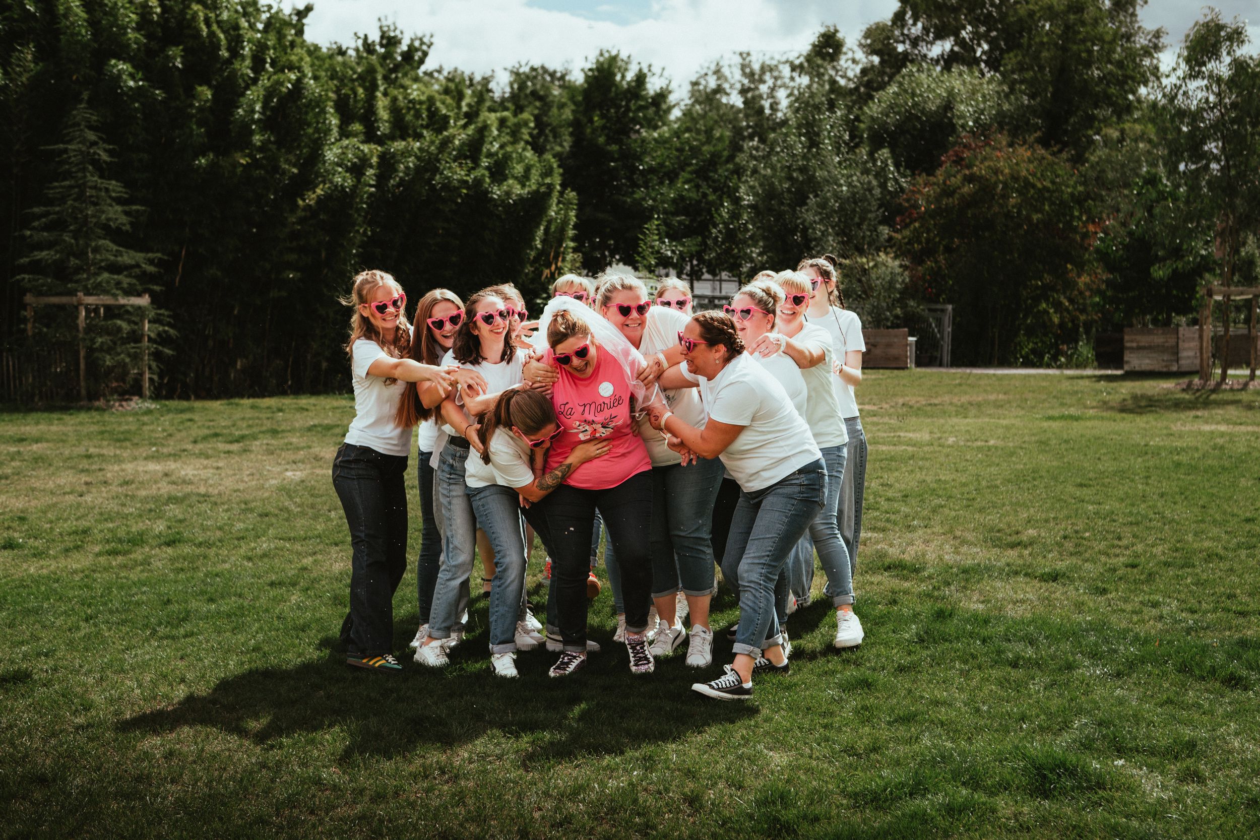 Group of women in white shirts hugging and celebrating outdoors on green grass with trees in background.