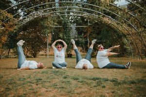 Four women sitting on grass under a metal arch trellis, playfully posing with legs in the air outdoors.